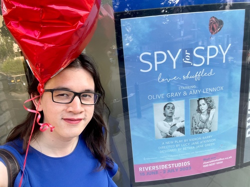 A selfie! I have light skin, dark brown hair, and rectangular glasses. I'm smiling at the camera, holding a heart-shaped balloon, and standing in front of one of the posters for the play.